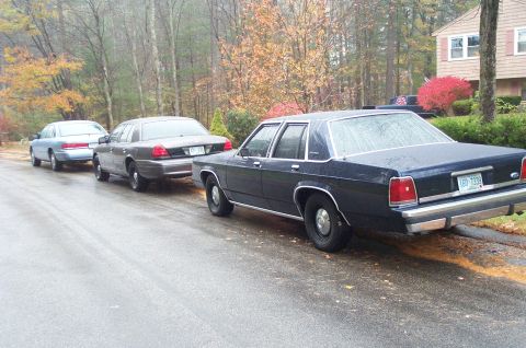 1991 Ford Crown Victoria LTD - Three sedans are parked on a wet street, with the closest one being a dark blue model.