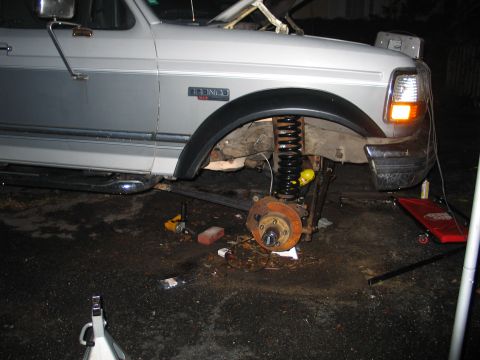 1992 Ford Bronco - Front passenger-side view of a silver Ford Bronco at night with the wheel removed, exposing the brake hub, rotor, and a black coil spring s…