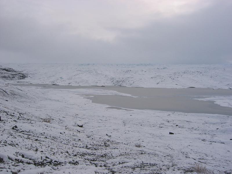 Snow-covered hills around a gray frozen lake under overcast sky