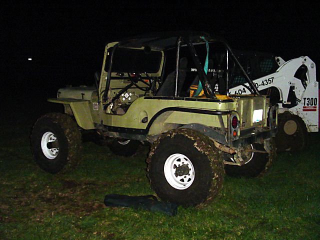 Olive green Jeep CJ-5 with a roll cage, mud-caked tires, and a white skid steer loader behind it