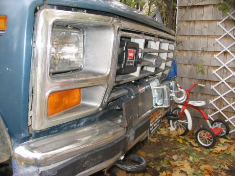1986 Ford Bronco - A winch is mounted on the front grille of a blue vehicle, with a child's tricycle parked to the side.