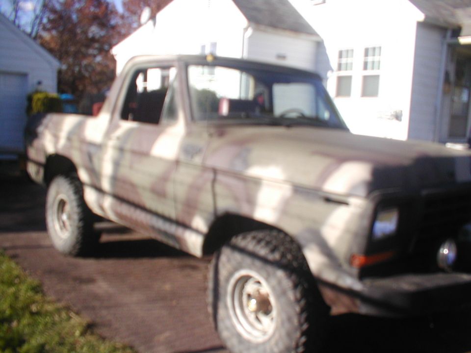 Lifted Ford Bronco in tan camo paint on mud tires and white spoke wheels beside a white house