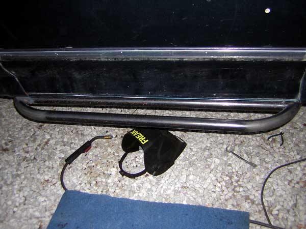 Black tube rock slider welded under a dark vehicle's rocker with welding leads and gloves on gravel