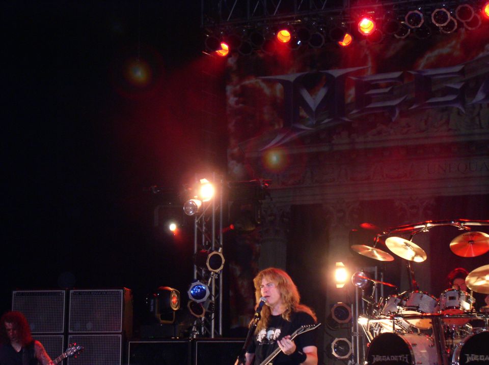 Musician with long blonde hair plays guitar and sings on stage with drums and speakers behind him