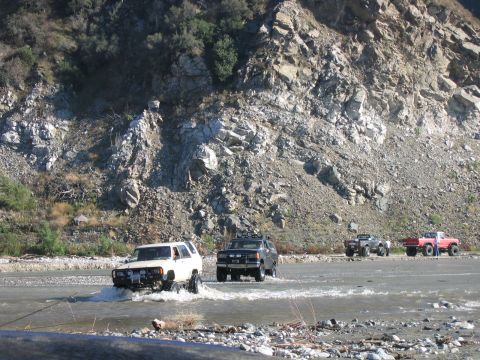 1992 Ford Bronco - A convoy of off-road vehicles navigates a riverbed, with the lead vehicle appearing to be a 1992 Ford Bronco.