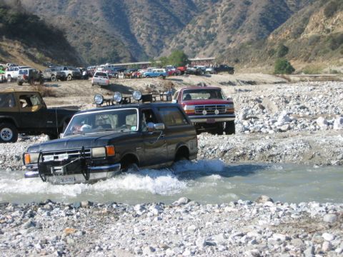 1985 Ford Bronco - Black Ford Bronco navigates a rocky riverbed, with other off-road vehicles visible in the background.