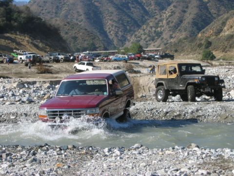 1985 Ford Bronco - Two off-road vehicles navigate a shallow river in a mountainous area with other vehicles parked on the bank.