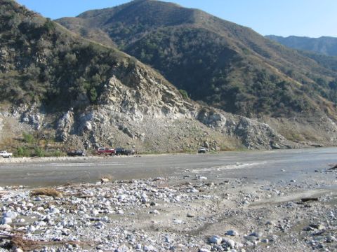 1985 Ford Bronco - Multiple vehicles are parked in a dry riverbed, with a mountain range visible behind them.