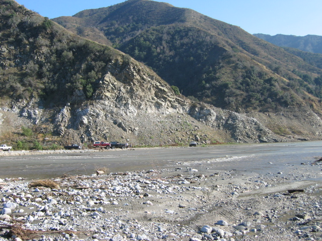 Distant vehicles parked on a gravel riverbank below a rocky mountainside