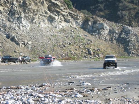 1985 Ford Bronco - Several off-road vehicles navigate a rocky riverbed under a steep, rocky hillside.