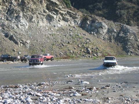 1985 Ford Bronco - Several trucks, including a 1985 Ford Bronco, navigate a shallow riverbed with a rocky cliff in the background.
