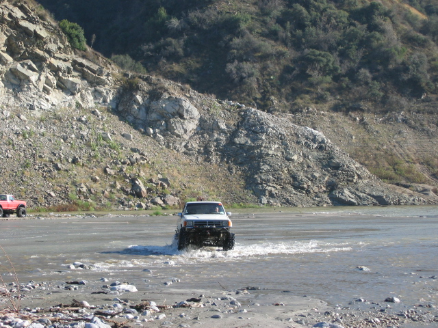 Lifted Ford truck fording a wide rocky river below a steep cut bank