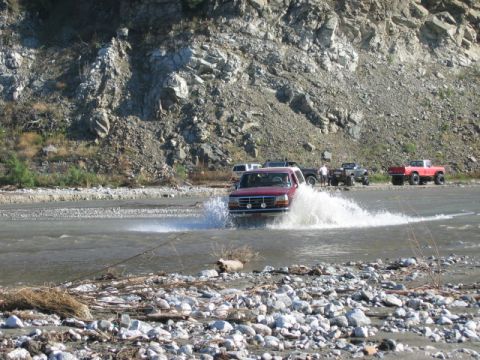 1985 Ford Bronco - Several trucks navigate a shallow river, kicking up water as they go.