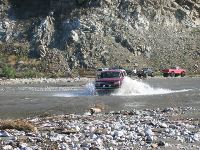 Red Ford Bronco driving through river splashing water, rocky hillside behind