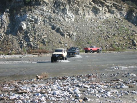 1985 Ford Bronco - Multiple off-road vehicles navigate a rocky riverbed, with a mountain range forming the backdrop.