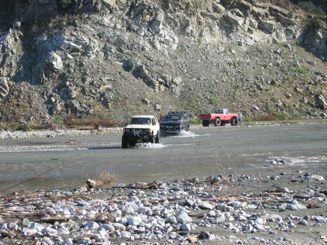 White SUV and other trucks crossing a shallow river in a rocky canyon