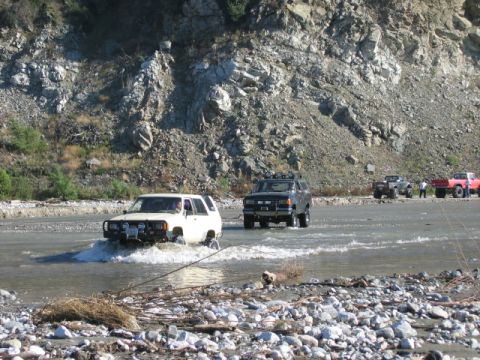 1985 Ford Bronco - A white 1985 Ford Bronco navigates a rocky riverbed, with other off-road vehicles following behind.