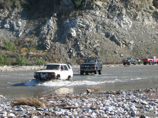 White and dark SUVs crossing a rocky river with vehicles parked on the far bank