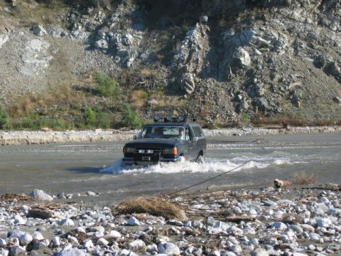 1985 Ford Bronco - Black Ford Bronco navigating a river crossing with rocky banks and a steep hillside.