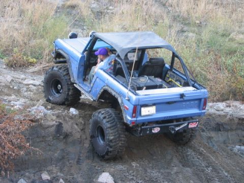 1985 Ford Bronco - Modified blue Ford Bronco navigating an off-road trail with large, aggressive tires.