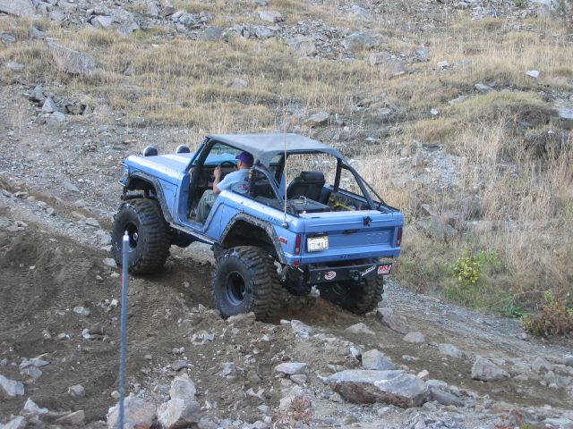 Blue Ford Bronco on large off-road tires climbing a rocky dirt hill, rear 3/4 view