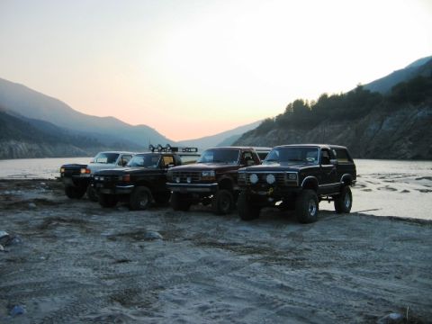 1985 Ford Bronco - Four SUVs are parked on a sandy shore next to a lake with mountains in the background.