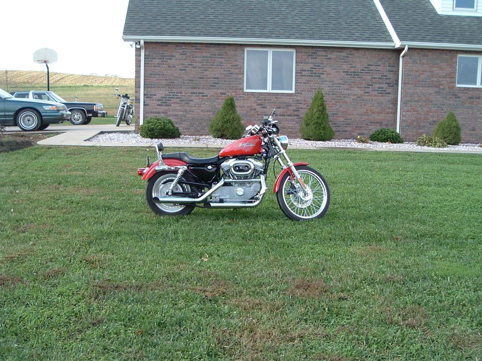 Red Harley-Davidson motorcycle parked on grass in front of a brick building; two cars and another motorcycle are visible in the background