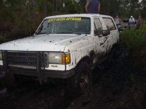 1989 Ford Bronco - This muddy white SUV is stuck in a swampy area with people around.