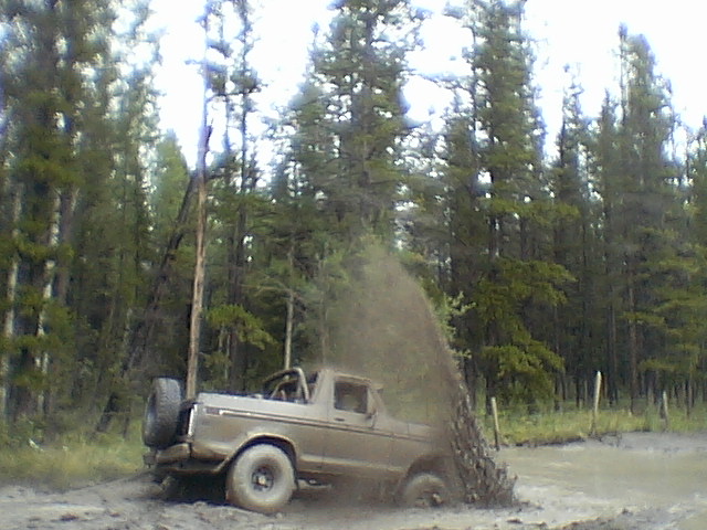 Mud-covered topless Ford Bronco throwing a rooster tail of mud in a bog surrounded by evergreen trees
