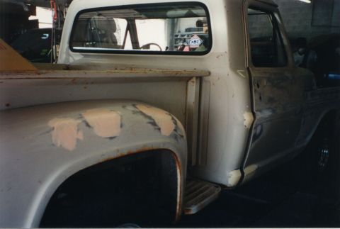 1968 Ford F-100 - Right-rear three-quarter view of a stepside pickup in bare primer during bodywork, showing tan body filler patches spotted across the rear…