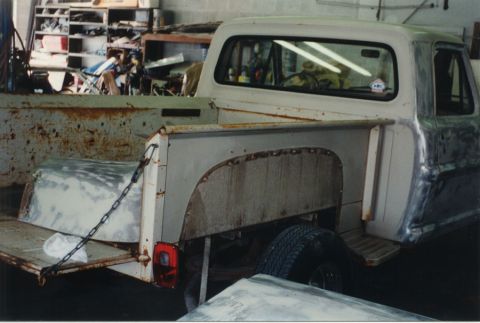 1968 Ford F-100 - Right-rear three-quarter view of a weathered classic pickup in a shop, its bed sides stripped to bare and rusty metal with the tailgate dow…