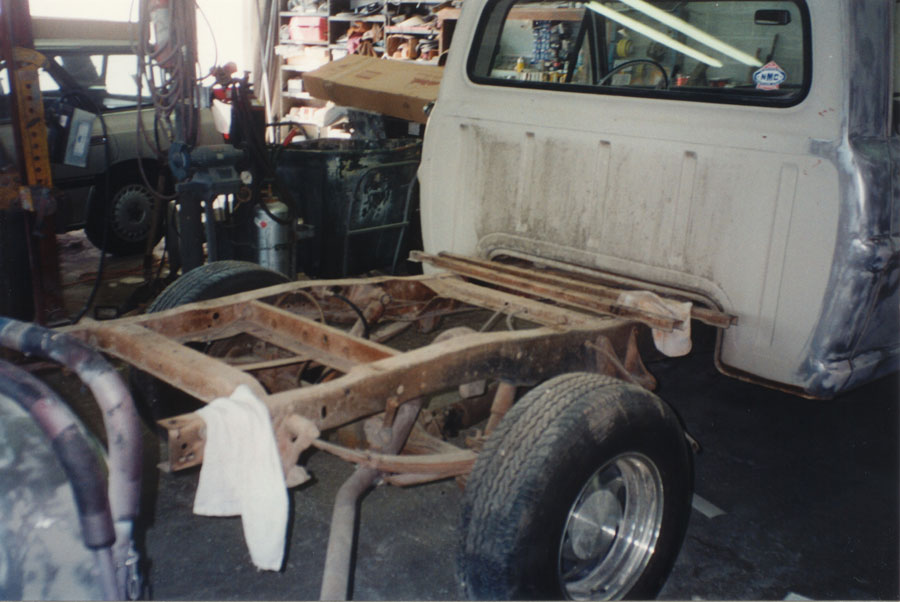 Pickup with bed removed showing bare rusty frame, leaf springs and driveshaft behind white cab
