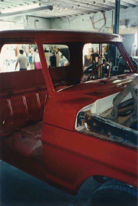 1968 Ford F-100 - Close-up of a classic Ford pickup cab in fresh bright red paint inside a shop, viewed from the right rear looking forward through the open…