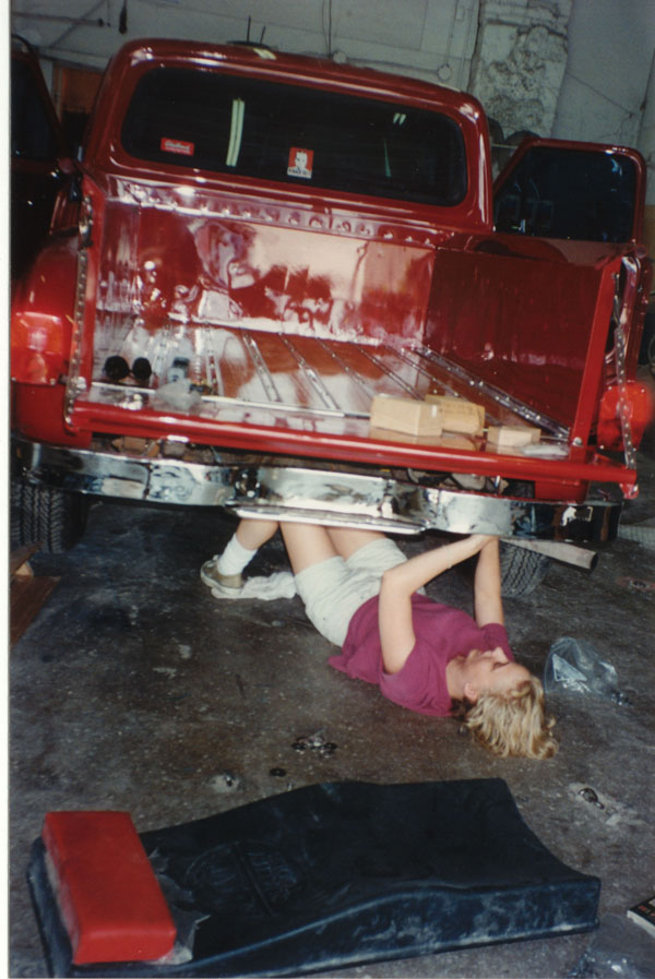 Person working under the rear bumper of a red classic Ford pickup in a garage