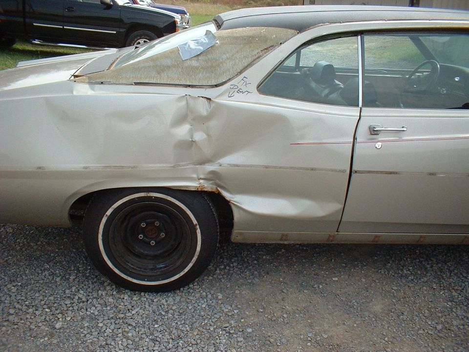 Dented gold rear quarter panel marked 'FIX DENT' on a two-door hardtop, parked on gravel