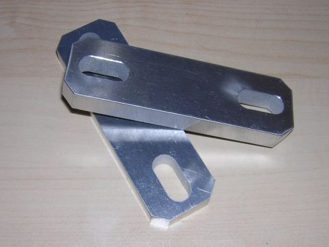 1998 Ford Crown Victoria - Two machined aluminum brackets with chamfered edges and slotted mounting holes, resting on a light wood surface.