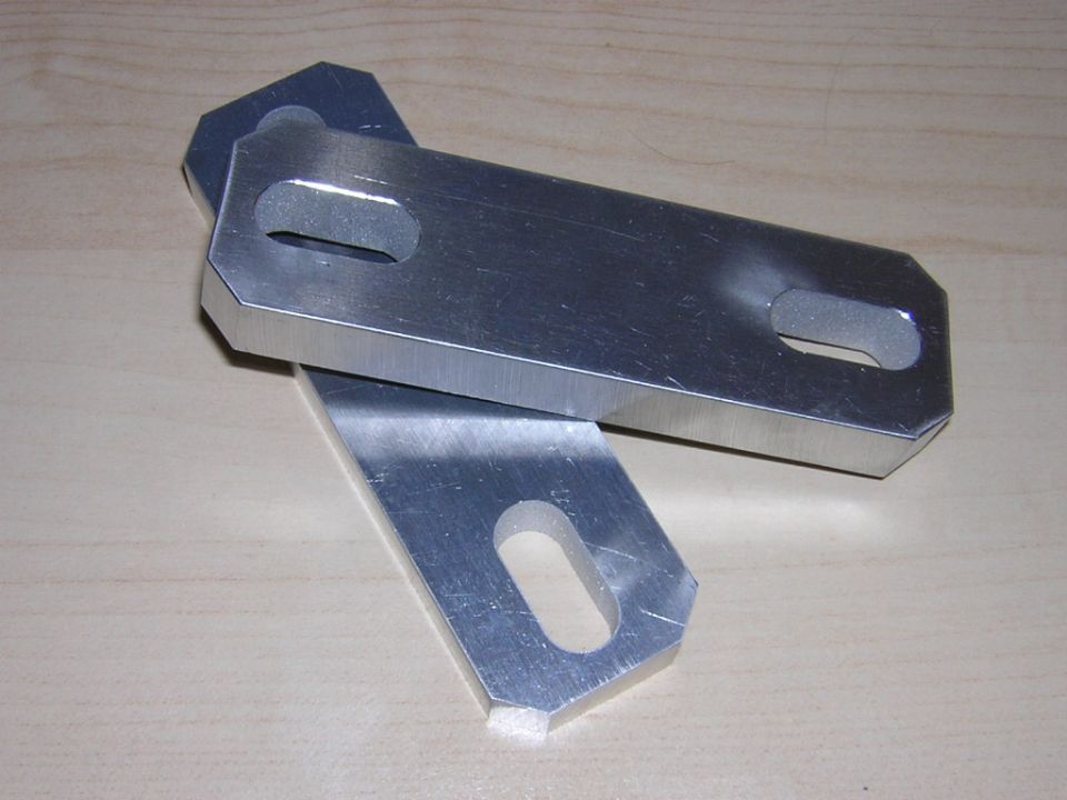 Two machined aluminum brackets with slotted holes lying on a wood surface