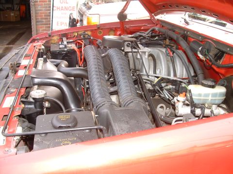 1994 Ford Bronco - Engine bay of a red truck seen with the hood up in a shop, showing a fuel-injected V8 with cast intake plenum, twin ribbed black air intake…