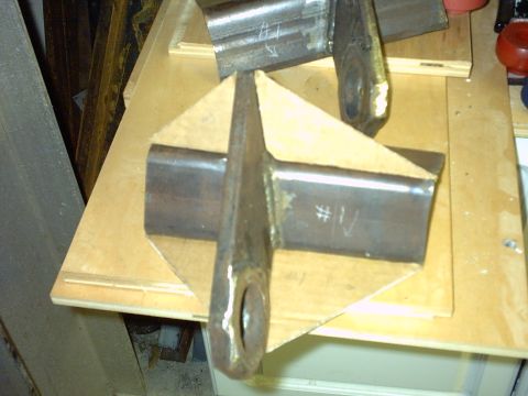 1995 Ford Bronco - Two custom-fabricated steel brackets on a plywood workbench, each made from a section of channel with a flat tab welded on that has a large…