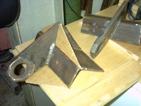 1995 Ford Bronco - A pair of shop-fabricated steel suspension brackets sitting on a plywood workbench, each built from welded plate with a large bushing bore…