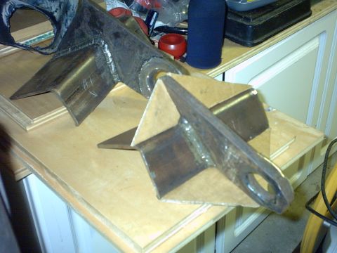 1995 Ford Bronco - Two shop-fabricated bare-steel suspension brackets sitting on wooden cabinet doors, each built from folded plate with gusseted webs and a w…