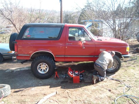 1987 Ford Bronco - A person works on the undercarriage of a red 1987 Ford Bronco, which has a spare tire mounted on the rear.