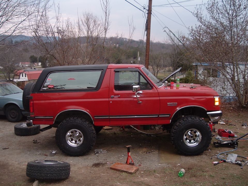 red 1987 Ford Bronco with lifted suspension and off-road tires, hood up, parked outside with tools around it
