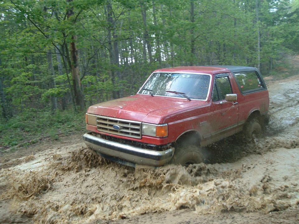 Red 1987 Ford Bronco driving through mud on a wooded trail