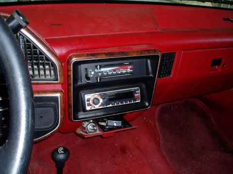 1990 Ford Bronco - Passenger-side view of a red vinyl truck dash with woodgrain center bezel, slide-lever heater controls above an aftermarket CD head unit, a…