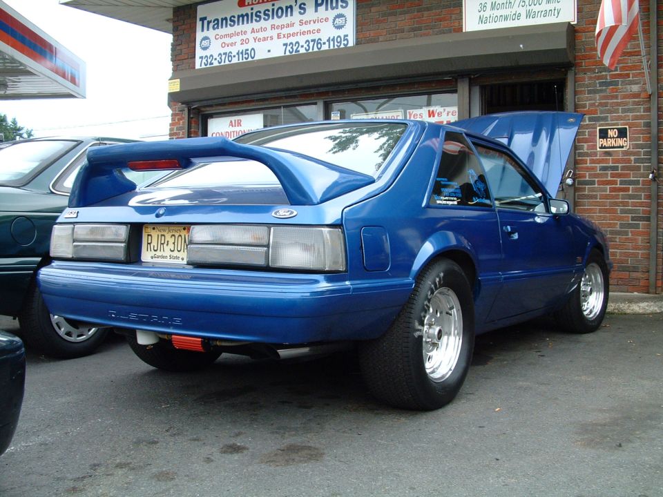 Blue 1988 Ford Mustang with a large rear spoiler and custom wheels parked outside a garage