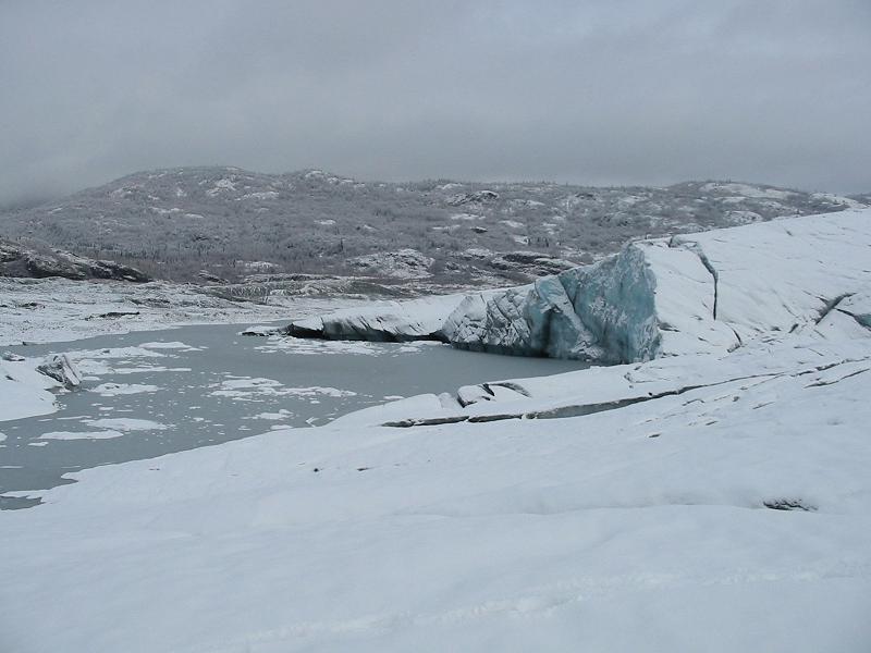 Glacier ice meeting a meltwater lake under an overcast sky in a snowy landscape
