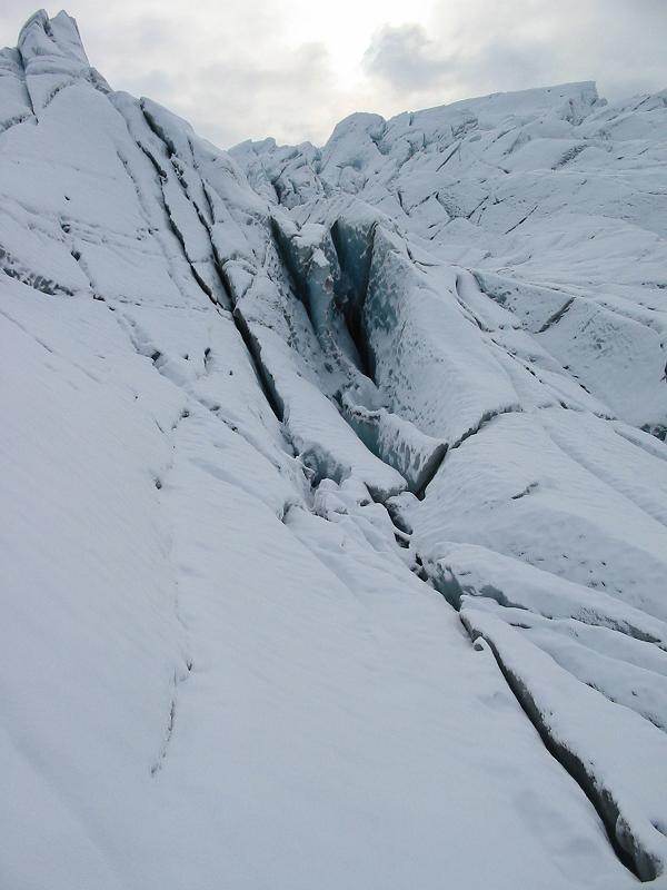 Snow-covered glacier with deep crevasses and exposed blue ice under cloudy sky