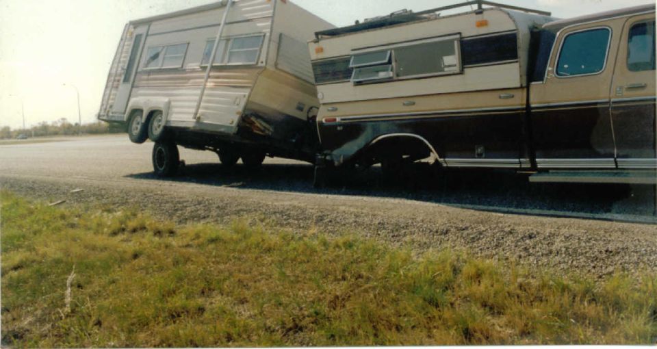 Brown and tan Ford pickup with camper shell hitched to a white travel trailer sagging at the hitch on a gravel shoulder