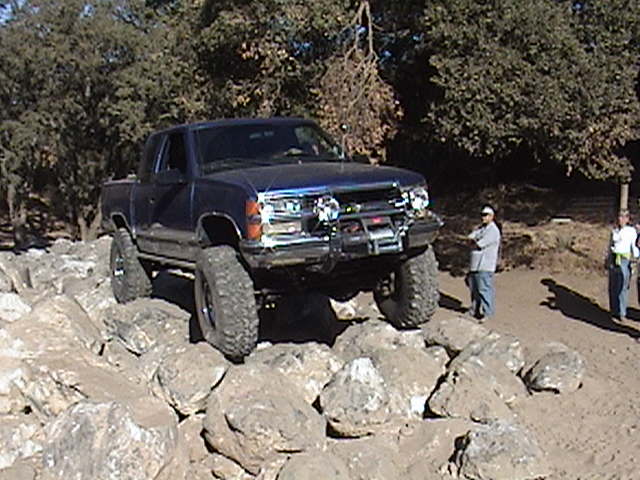 Blue pickup truck with large tires navigating a rocky trail. Two people stand nearby